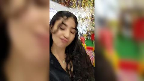 Media: A blurred video of a young woman with dark curly hair, closed eyes, and a serene expression, standing in a festive, colorful background with blurred, bright lights and decorations.