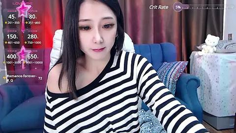 Media: Video of a young Asian woman with straight black hair, wearing a black-and-white striped top, sitting in a cozy room with a blue couch, teddy bear, and patterned cushions.