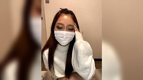 Media: Video of an Asian woman with long, straight dark hair, wearing a white mask, long-sleeved white top, and a white shirt. She appears to be sitting, with a blurred person in the background. The setting is indoors, with beige walls and a wooden floor.
