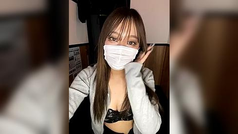 Media: Video of a young Asian woman with long brown hair, wearing a white surgical mask, black lace bra, and grey cardigan, seated in a dimly lit restaurant, blurred faces in the background.