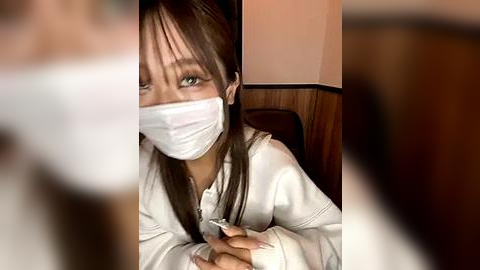 Media: Video of a young Asian woman with long brown hair and green eyes, wearing a white face mask, sitting in a restaurant. She has a sad expression.