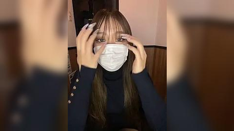 Media: Video of a young Asian woman with long, straight brown hair, wearing a black turtleneck, white mask, and gloves, adjusting her mask while seated indoors with blurred, wooden paneling background.