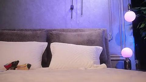 Media: A video of a modern bedroom featuring a plush, gray upholstered headboard, two white pillows, and a small book on a neatly made bed. The background showcases a textured, light blue wall with two pink, spherical pendant lights and a plant on the right.