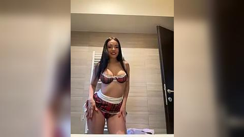 Media: A video of a young woman with long black hair, fair skin, and medium breasts, wearing a plaid bikini, standing in a modern bathroom with beige tiled walls and a towel rack.