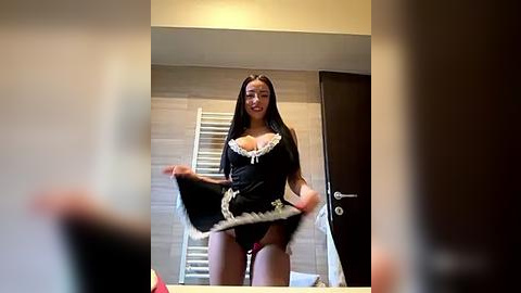 Media: Video of a young woman with long dark hair in a black maid outfit, lifting her skirt, revealing red panties, standing in a modern bathroom with beige tiles and a dark door.