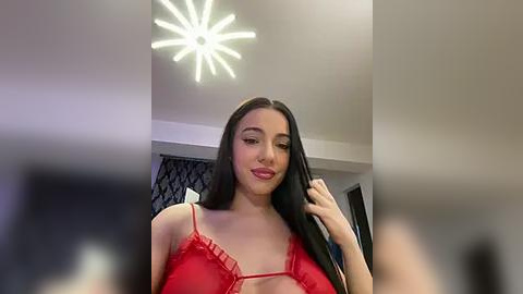 Media: Video of a young woman with long black hair, wearing a red lace bralette, standing in a modern room with a chandelier.