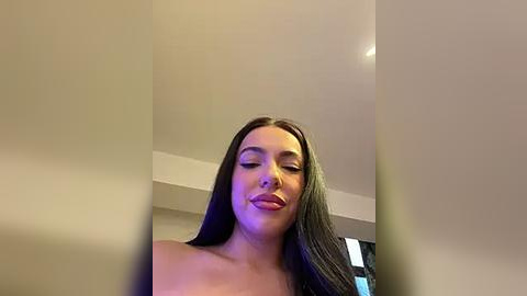 Media: A video of a young woman with long, straight black hair and fair skin, wearing a strapless top, making a pouty face. The background is a simple, beige-colored room with a ceiling light.