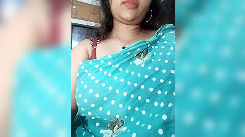 Media: A video of a South Asian woman with medium skin tone, wearing a turquoise polka-dot saree with embroidered floral patterns. She has long, wavy black hair and is indoors, possibly in a kitchen with cabinets in the background.