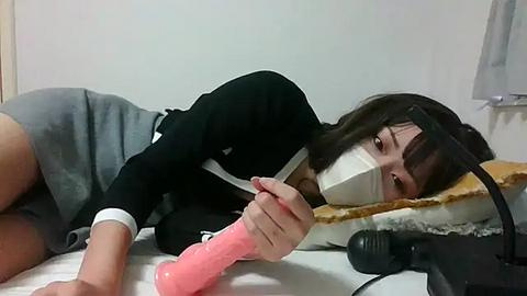 Media: Video of an Asian woman with straight black hair, wearing a black sweater and gray shorts, lying on a bed with a pink dildo and a vibrator, gagged with a white cloth.