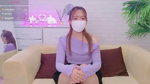 Media: Video of an Asian woman with long red hair, wearing a purple off-shoulder top, white face mask, and black skirt, sitting on a beige sofa with brown pillows, in a modern room with white brick wall, potted plant, and a glowing neon sign.