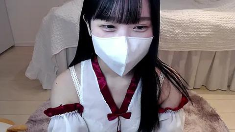 Media: Video of an Asian woman with long black hair, wearing a white mask, off-shoulder white dress with red trim, kneeling on a beige floor, in a room with white furniture and a bed.