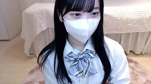 Media: Video of a young Asian woman with long black hair, wearing a white school uniform with a plaid bow tie, a face mask, and sitting on a pink fur rug in a simple, beige-lit room.