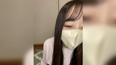 Media: Video of an Asian woman with long black hair, wearing a beige face mask, white top, and sitting on a patterned rug in a room with beige walls.