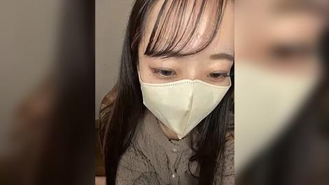 Media: A close-up video of an Asian woman with long black hair, wearing a beige face mask, looking downward with a serious expression.