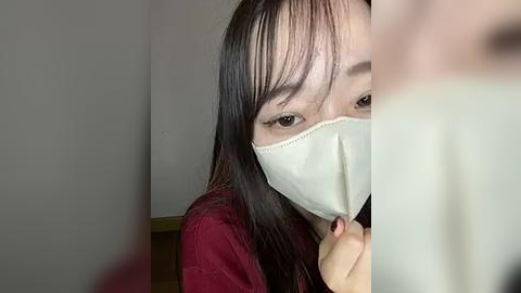 Media: Video of an East Asian woman with long black hair, wearing a white surgical mask, standing indoors against a plain, light-colored wall, partially obscured by another person's hand.