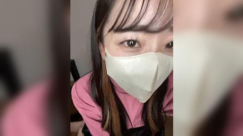 Media: Video of an Asian woman with long, straight brown hair, wearing a white face mask and pink shirt, gazing directly at the camera, blurred background.