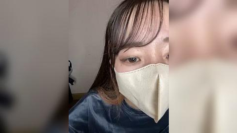 Media: Video of a young woman with straight, shoulder-length brown hair, partially covered by a beige face mask, wearing a blue surgical gown. She has fair skin and is looking directly at the camera. The background is blurred and neutral-toned.