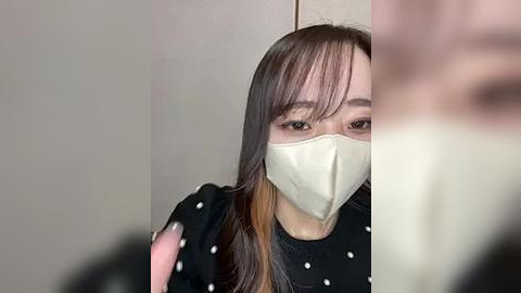 Media: Video of an East Asian woman with long straight brown hair, wearing a beige mask, black polka-dotted top, and gloves, obscured by a blurred figure in the background.