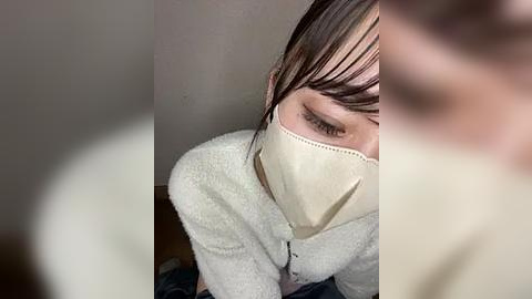 Media: Video of a person wearing a beige face mask, white long-sleeve shirt, and dark hair, crouched in a dimly lit room with blurred background.