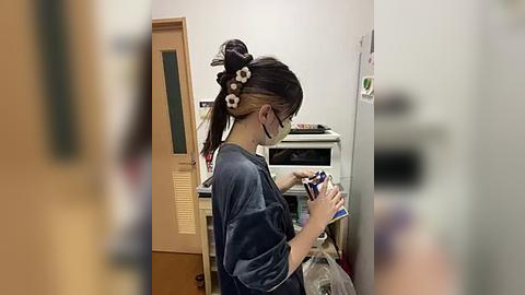 Media: Video of an Asian woman with black hair styled in a high bun, wearing glasses and a blue top, using a phone in a dimly lit room with a fridge and wooden cabinet.