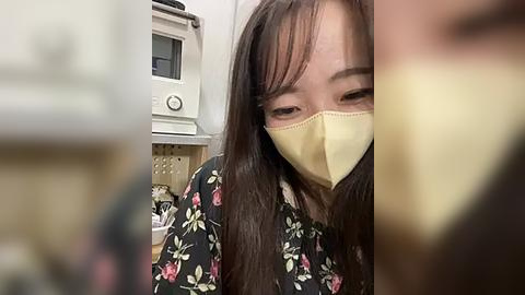 Media: Video of an Asian woman with long brown hair, wearing a yellow surgical mask, black floral kimono, and sitting indoors near a white appliance, possibly a microwave, with a blurred background.