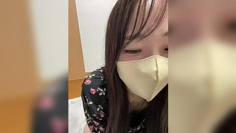 Media: Video of an Asian woman with long dark hair, wearing a yellow face mask and floral-patterned black dress, leaning forward, blurred background.