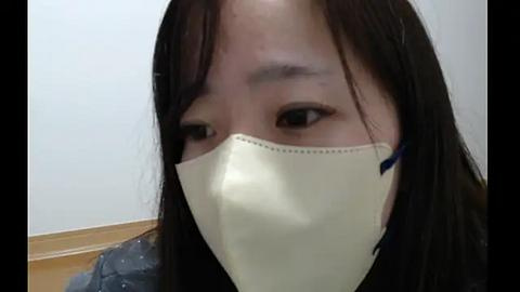 Media: Video of a young woman with straight black hair, wearing a beige surgical mask, looking down, in a plain, light-colored room with a wooden floor.