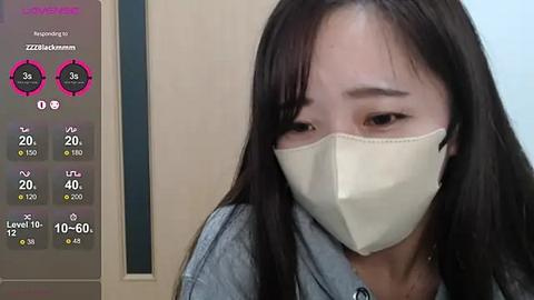 Media: Video of an Asian woman with long black hair, wearing a beige face mask and light denim jacket, in a room with a digital thermometer showing 25\u00b0C.