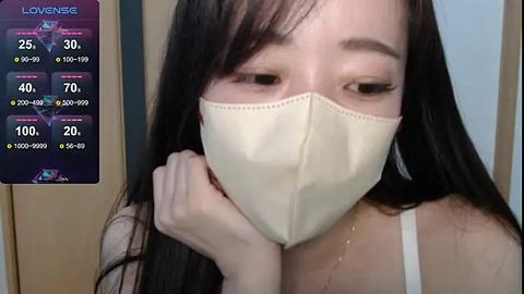 Media: Video of an Asian woman with long black hair, wearing a white face mask, looking down. Background shows a smartphone screen with a \"Love & Live\" app displaying her stats.