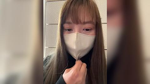 Media: Video of a young woman with straight, light brown hair and bangs wearing a white face mask, black jacket, and a black shirt, standing in front of a white, perforated wall.