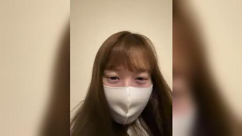 Media: Video of a young Asian girl with long brown hair and bangs, wearing a white surgical mask, looking down, against a beige wall.