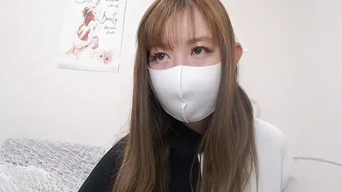 Media: Video of an Asian woman with long, straight brown hair, wearing a white mask, black top, and white jacket, in a minimalistic, white-walled room with a pink poster and gray textured background.