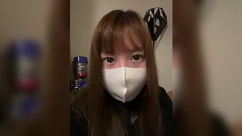 Media: Video of a young Asian woman with long brown hair, wearing a white face mask, black fur-lined jacket, and standing in a dimly lit room with abstract art on the wall.