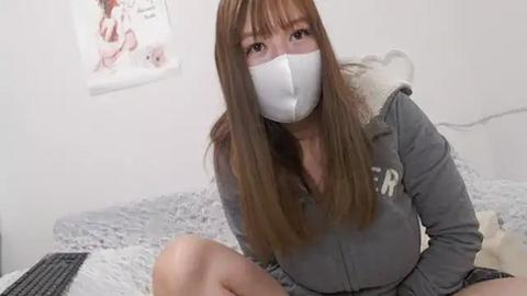 Media: Video of a young Asian woman with long, straight brown hair, wearing a white face mask, gray hoodie, and black shorts, sitting on a bed with a gray blanket in a minimalistic bedroom.