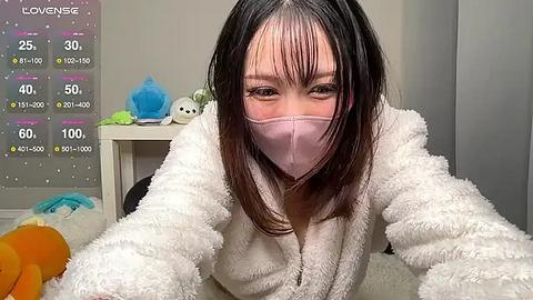 Media: Video of an Asian woman with long black hair wearing a pink face mask, a white fuzzy robe, and a blue plush toy in a cozy room.