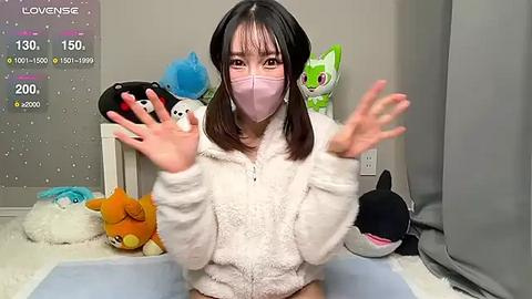 Media: A video of a young Asian woman with long black hair, wearing a pink face mask, white fuzzy hoodie, and holding a blue plush toy, surrounded by various plush toys in a cozy room.