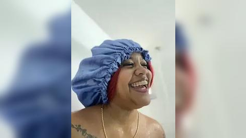 Media: A video of a smiling, topless, medium-skinned Black woman with red hair wrapped in a blue shower cap, wearing a gold necklace. Background is blurred and white.