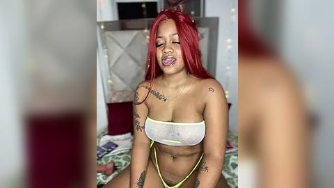 Media: Video of a young, light-skinned Black woman with bright red hair, wearing a sheer white bandeau top and yellow thong, with numerous tattoos, lying on a bed with colorful sheets, in a dimly lit bedroom.