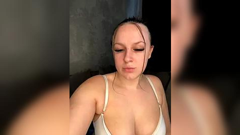 Media: A video of a young Caucasian woman with light skin and medium-length dark hair, tied back, wearing a white tank top, sitting in a dimly lit, concrete room.