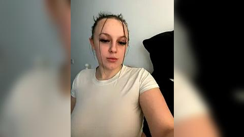 Media: A video of a young Caucasian woman with fair skin, brown hair tied back, wearing a tight, white, short-sleeved shirt that accentuates her small breasts. She appears to be indoors with a black cushion in the background.