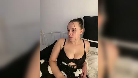 Media: Video of a fair-skinned woman with dark hair in a bun, wearing a black lace bra and matching skirt, sitting on a bed with white pillows and a black-and-white floral-patterned blanket, in a dimly lit room.