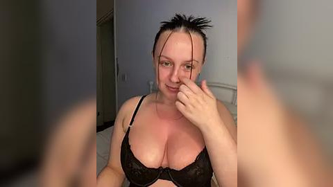 Media: A video of a fair-skinned woman with short dark hair, wearing a black lace bra, sitting on a bed. She has a playful expression, touching her cheek with her finger.