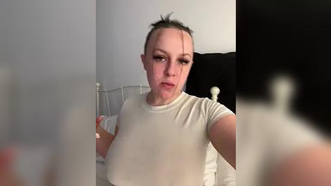 Media: Video of a Caucasian woman with fair skin and dark hair in a messy bun, wearing a white t-shirt. She appears to be taking a selfie in a dimly lit bedroom with a white metal bed frame and black pillow.