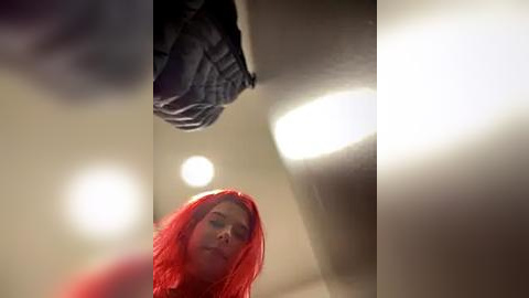 Media: Video of a woman with vibrant red hair, wearing a black jacket, standing in a dimly lit, modern room with soft, ambient lighting and beige walls.