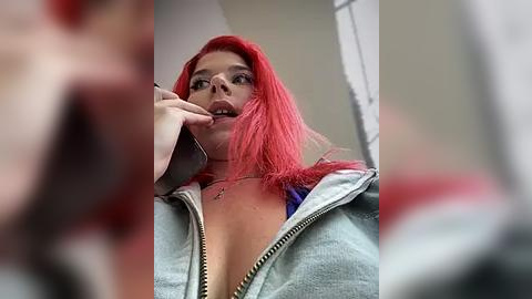 Media: Video of a young woman with vibrant pink hair, wearing a low-cut gray hoodie, talking on a smartphone while indoors with blurred background.