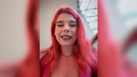 Media: Video of a young woman with vibrant pink hair, wearing a pink jacket, smiling with braces, in a modern indoor setting with large windows.