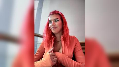 Media: Video of a fair-skinned woman with long, bright red hair, wearing a coral-orange top, smiling, in a modern indoor setting.