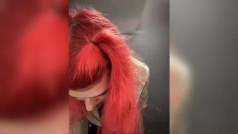 Media: Video of a person with bright red hair styled in a high ponytail, wearing a beige jacket, captured from a low angle, blurred background.