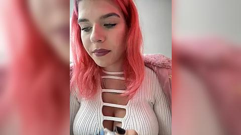 Media: Video of a young woman with vibrant pink hair, wearing a white, ribbed, crisscross top, sitting indoors, with a blurred pink background.