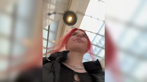 Media: Video of a young woman with red hair, wearing a black jacket, looking upward in a modern, glass-enclosed space with large windows and a pendant light.